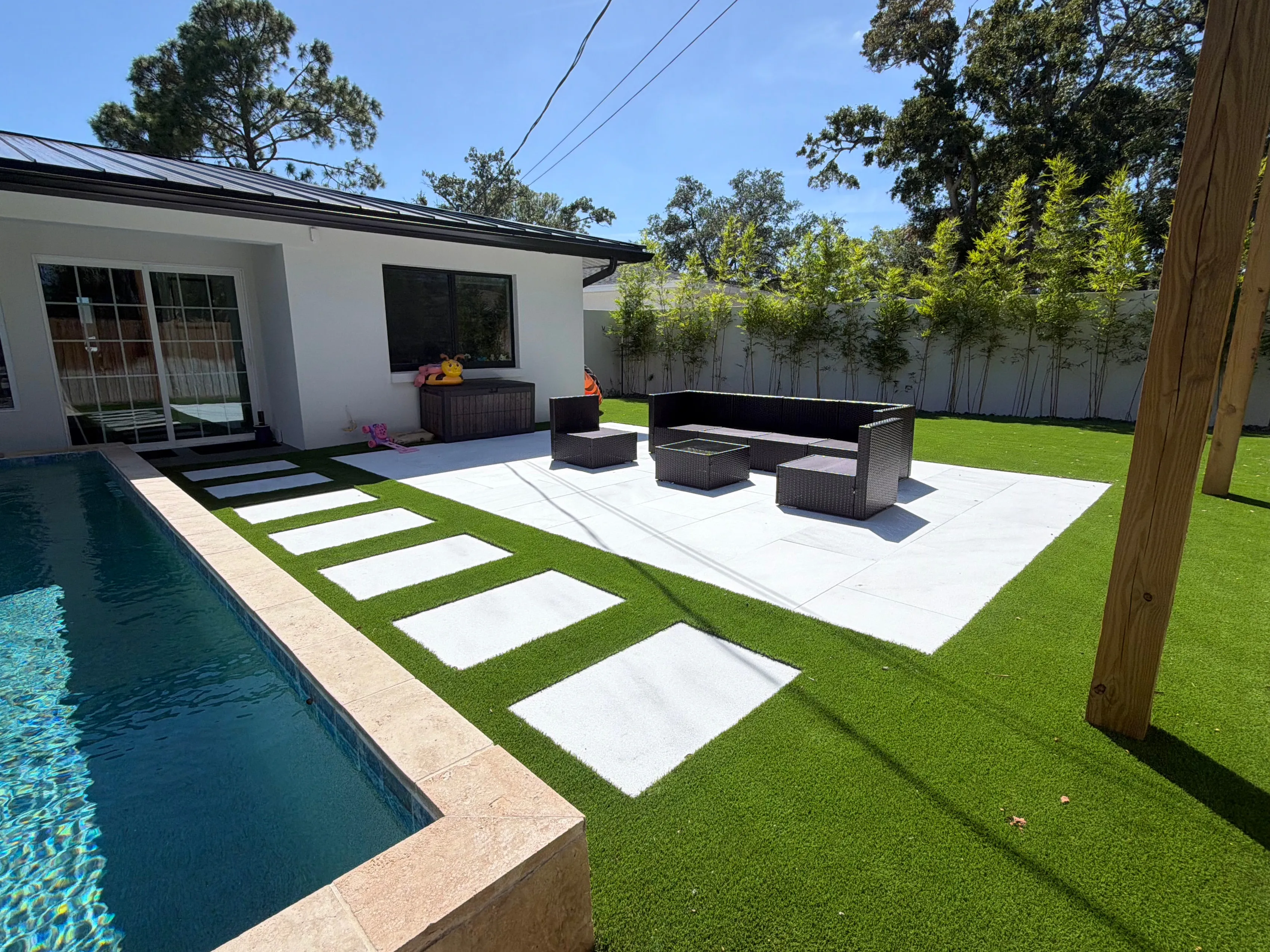 Artificial turf between white paver steppers beside a pool, with a patio seating area and bamboo along the fence