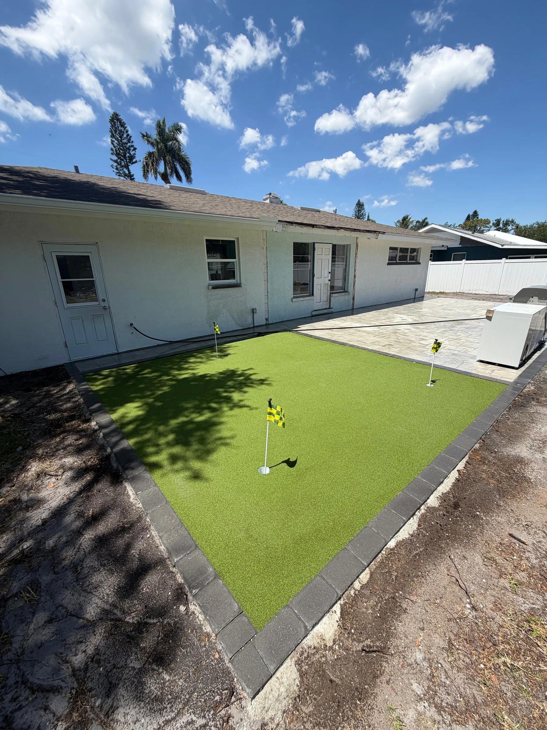 Backyard putting green with flags beside a white house and a paver border